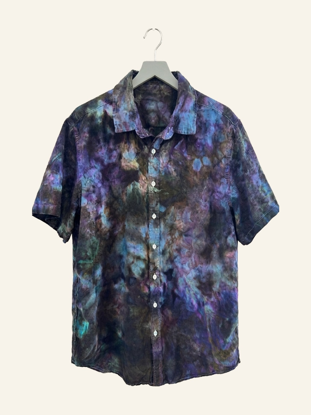 Hand ice-dyed cotton short-sleeve shirt in deep teal, violet, and slate on a pale grey hanger